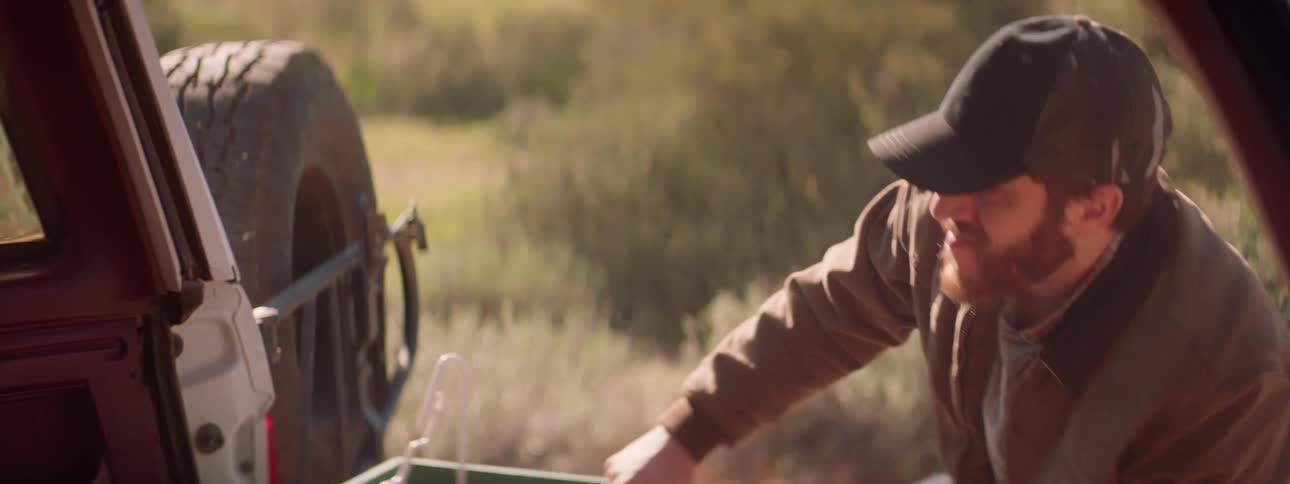 A man with a hat looking at the back of a truck with grass in the background