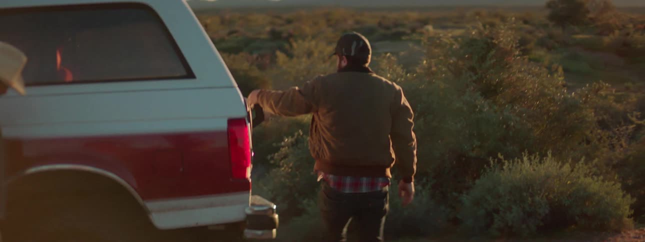 Two men stand next to a parked car in a rural area at sunset