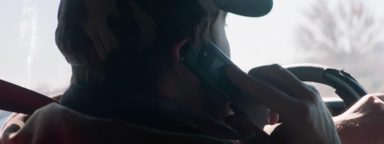 Man with beard and a cap talks on the phone while driving a car