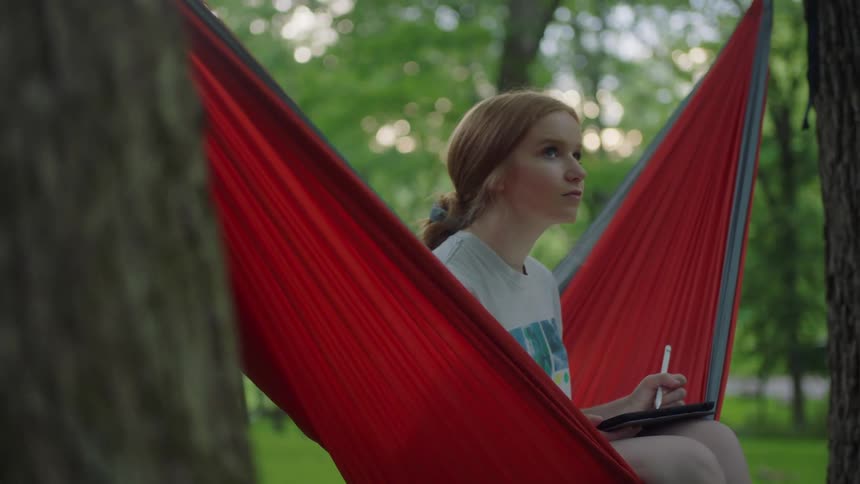A teenager sits in a red hammock in a park and writes about what she sees around her