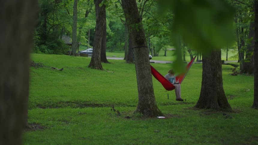 A young girl sits on a red hammock attached to trees in a park