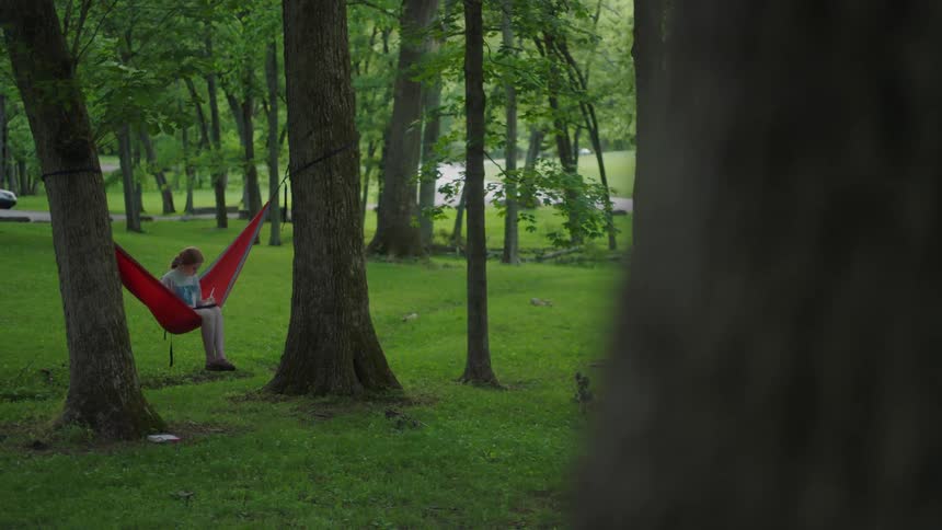 Girl with red hair and sits in a red hammock and uses a tablet in the forest
