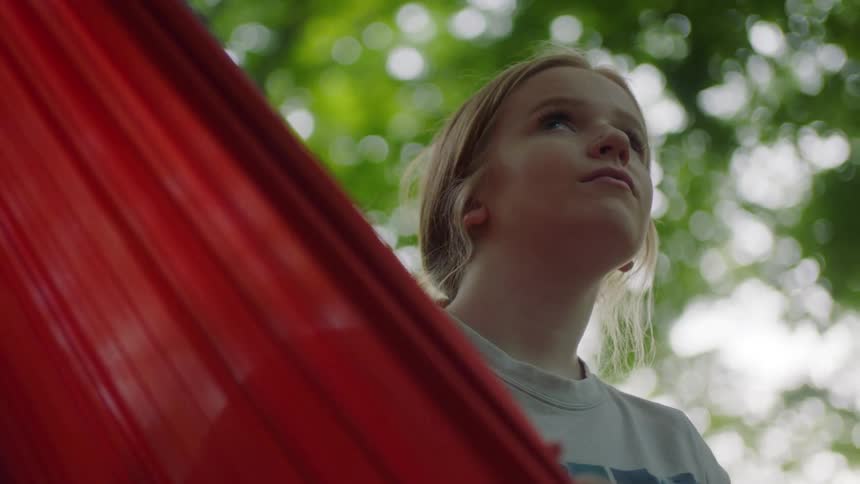 Girl with red hair and a grey long sleeve t shirt sits in a red hammock in the forest