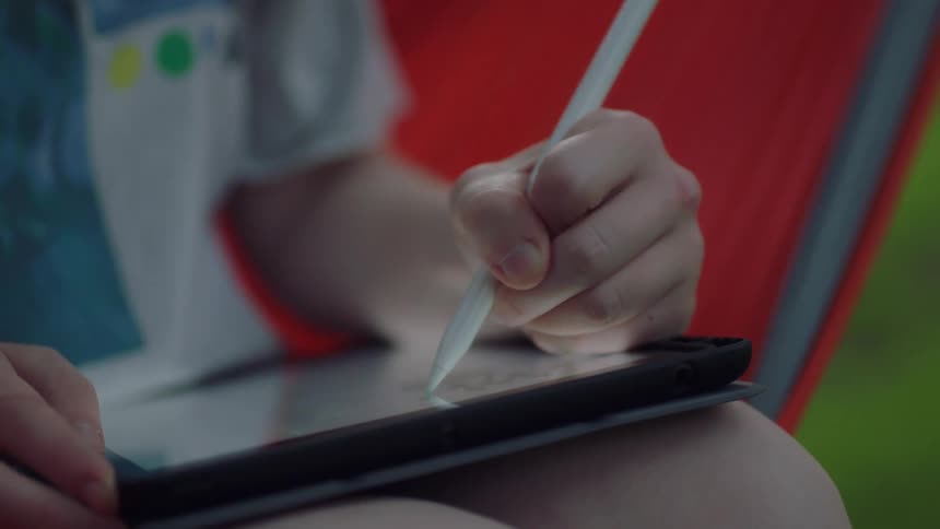 A young woman draws on a tablet using a stylus while sitting on a hammock outdoors