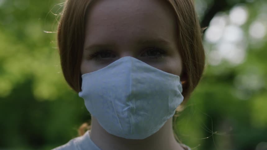 Girl with red hair and a light blue face mask looks at the camera in a forest