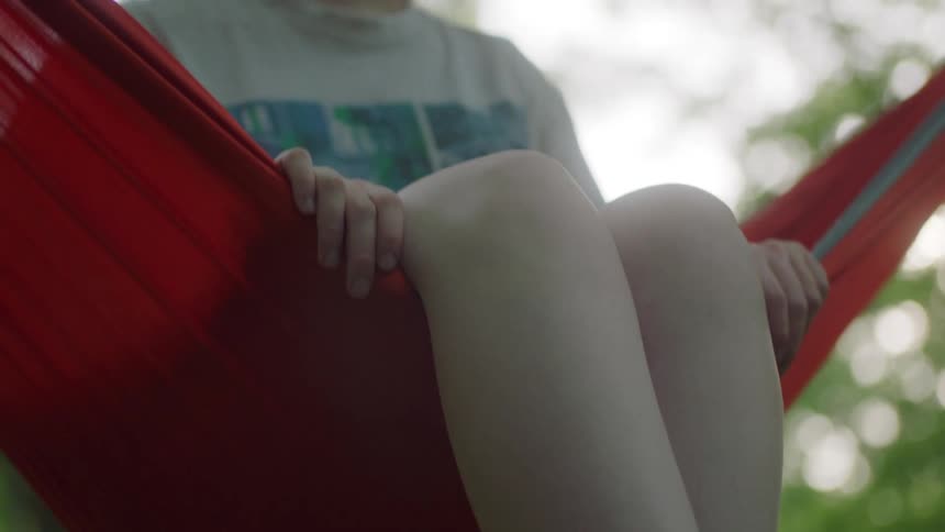 Girl with red hair brown sandals and shorts sits in a red hammock in the forest