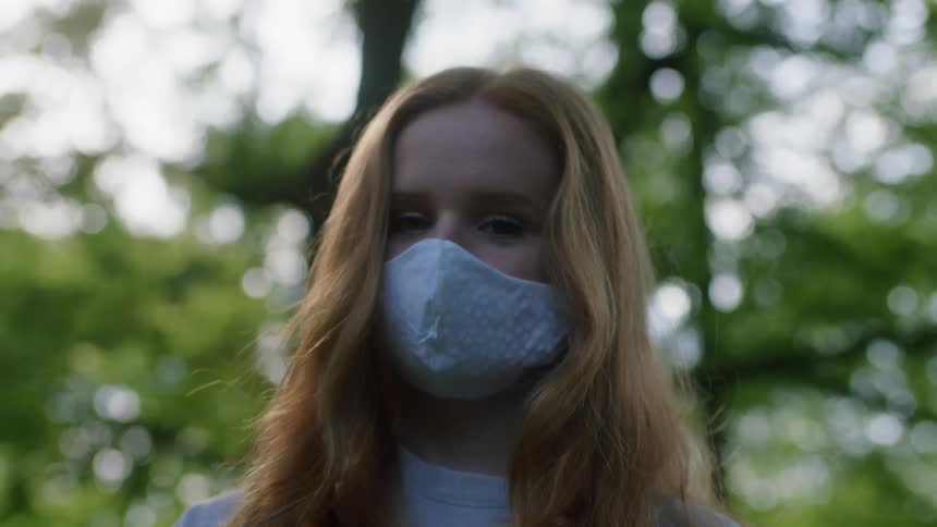 A young woman standing outdoors in a face mask looks around the area before looking into the camera