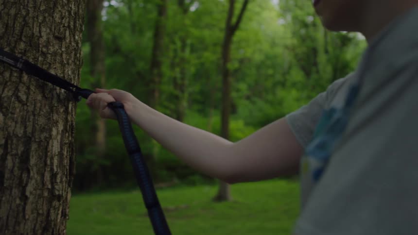 Girl with red hair a grey t shirt and shorts sets up a hammock with straps in the forest