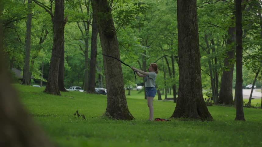 Girl with red hair a grey t shirt and shorts sets up a hammock with straps in the forest