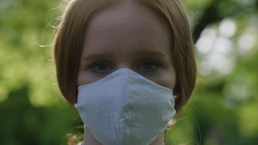 Girl with red hair and a light blue face mask looks at the camera in a forest