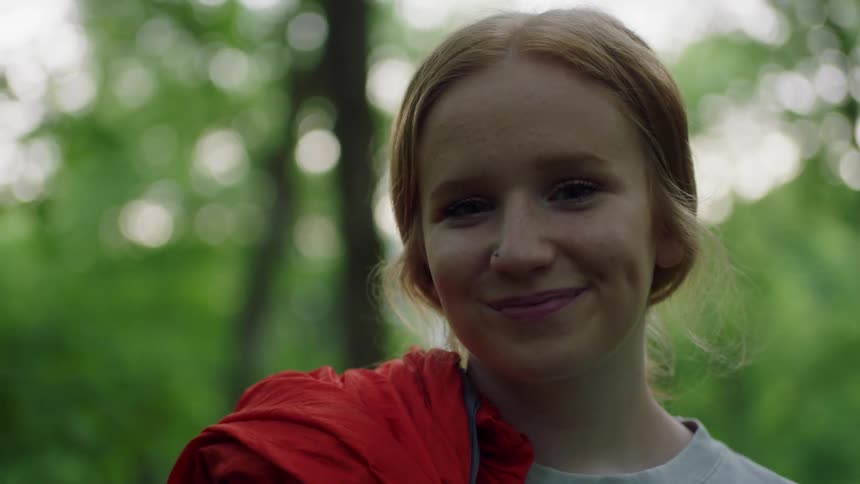 Woman with a red garment on her shoulder smiles while she is near a forest