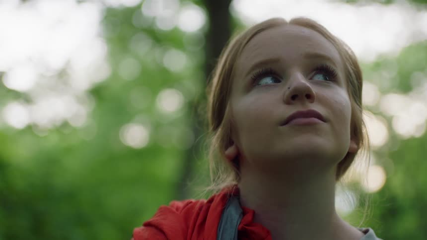 Girl with red hair looks at the tall trees and turns her gaze to the camera in a forest