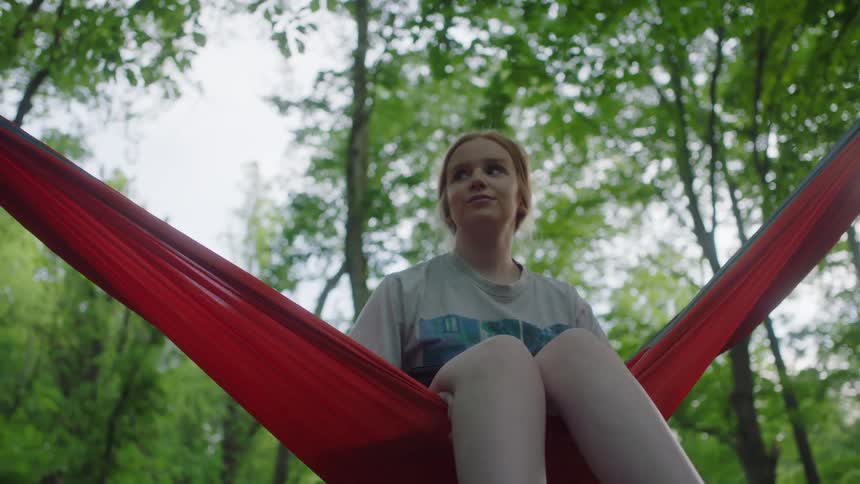 Girl with red hair sits in a red hammock in the forest