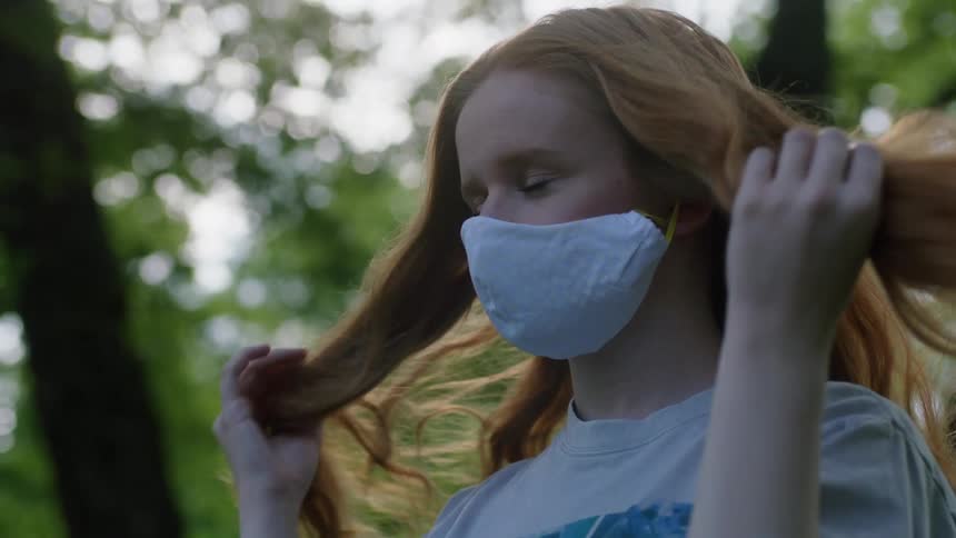 Red haired woman wearing face mask stands outdoors finger combing her hair