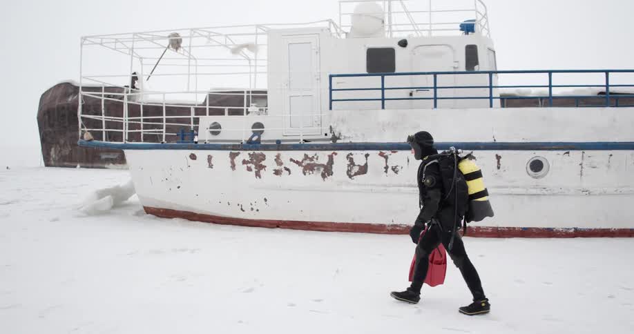 Person wearing scuba gear walks through lightly falling snow across packed ice past large boat