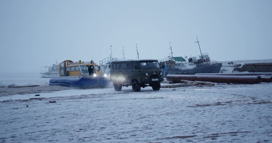 Bus towing an hovercraft out of the water onto the snow covered sands of a beach at winter