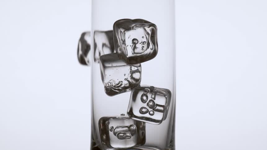 Ice cubes fall into a tall glass in front of a white background