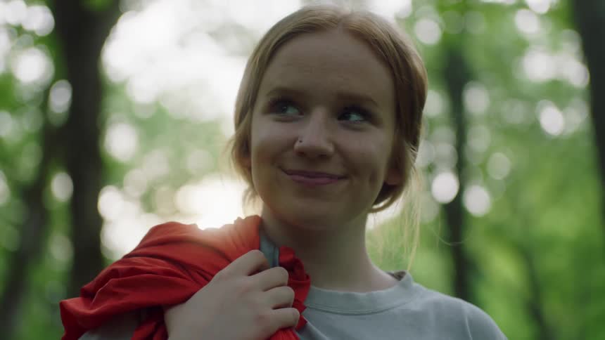 Girl with red hair looking at the camera holds a red hammock to set up in the forest