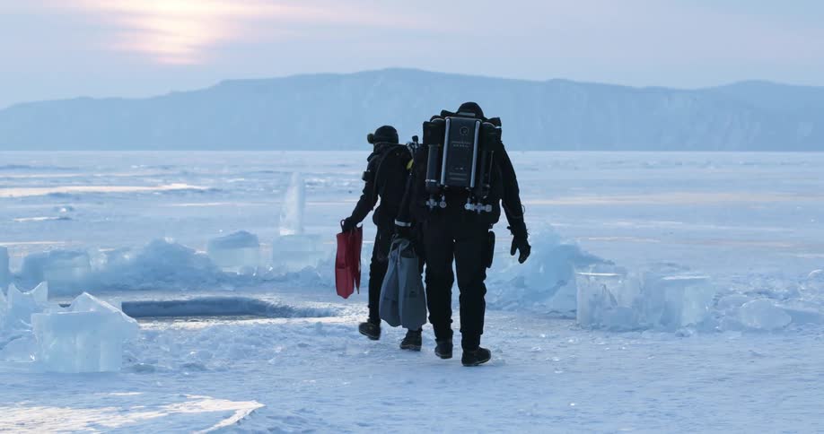 Two people with oxygen tanks and goggles holding flippers and walking on tundra ice