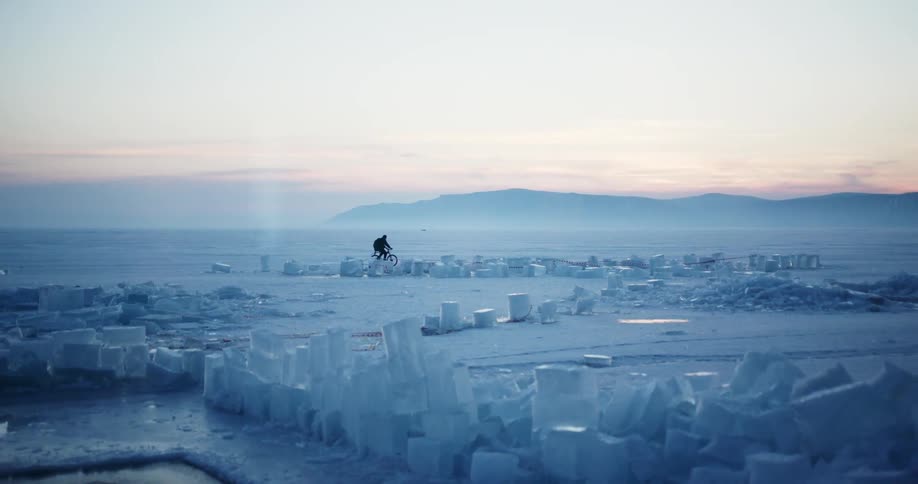 Bicyclists crosses white plain past various sized cubes and rectangle as red sky tops mountains
