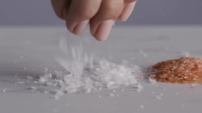 Person drops white salt crystals onto pale gray surface next to rust colored salt crystals