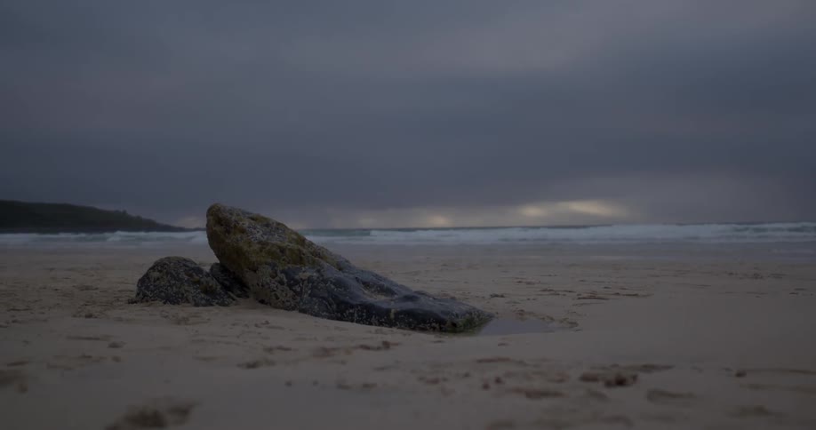 A few large rocks sit in the sand by the shore of a beach at sunset