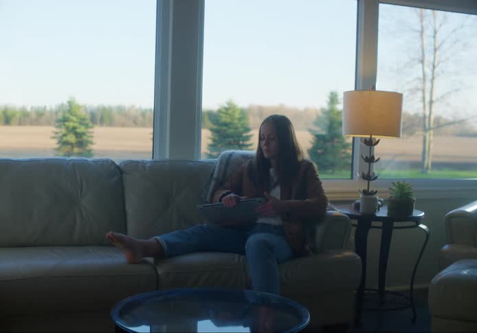 Woman with brown straight hair and jeans carrying a laptop and a cup of coffee sits down on a couch in the living room