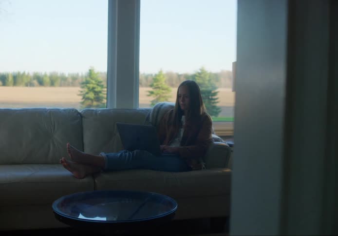 Woman sits on couch working on laptop in front of windows showing forest and fields