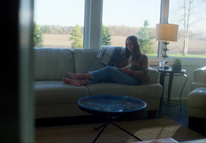 Woman with brown straight hair and jeans sits on a couch and writes in a notebook in the living room at home