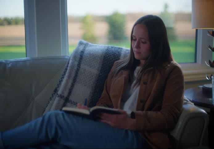Woman with brown straight hair and a brown jacket sits on a sofa and writes in a notebook in the living room at home