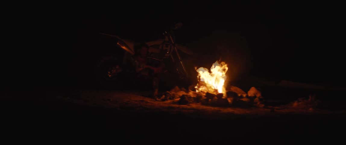 Man sits near motorcycle at night warmed by roaring campfire
