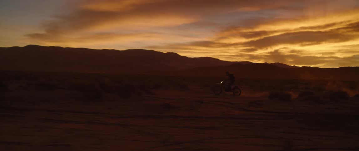 Horizon over mountains remains tinted orange and motorcyclist rides bike though scrub kicking up dust