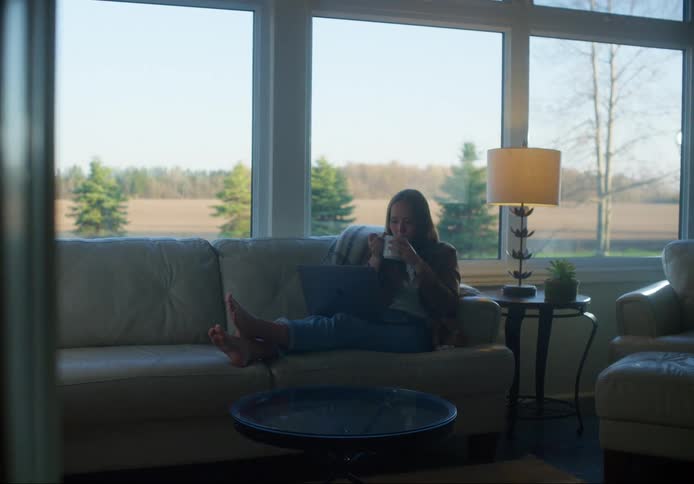 A woman sits on a couch with her laptop near some picture windows