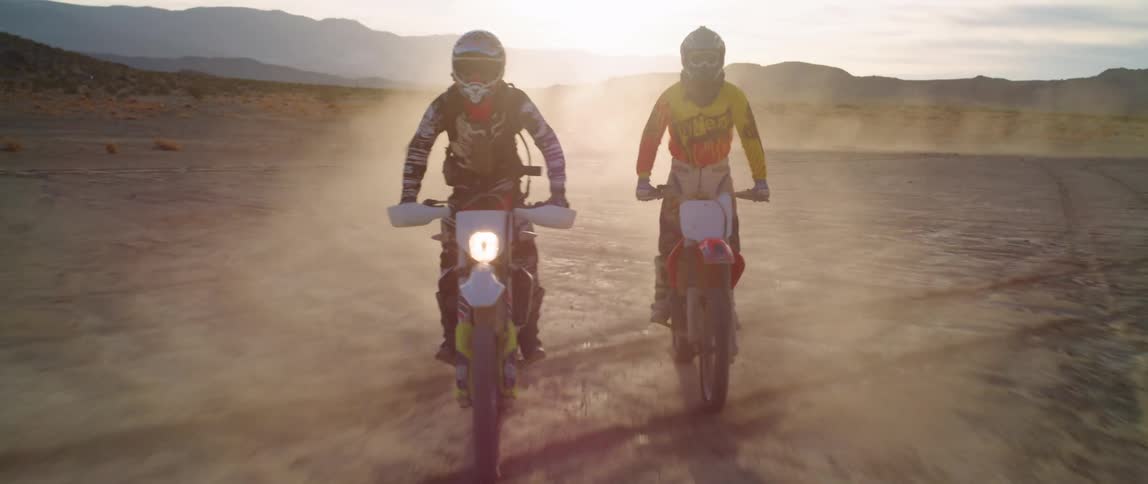 Two people ride their dirt bikes on the dusty field