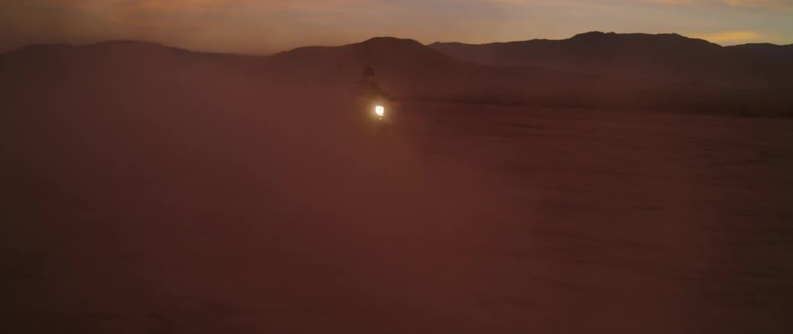 Motorcyclists rides across desert through dust with mountains on horizon and his headlight shining through the dust cloud