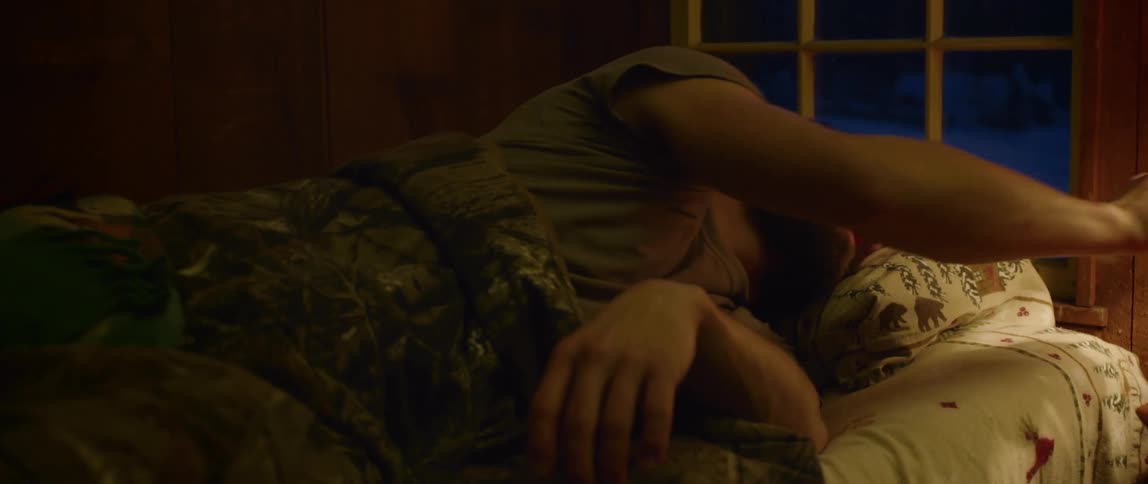 A bearded man laying in bed reaches over and turns on a lamp