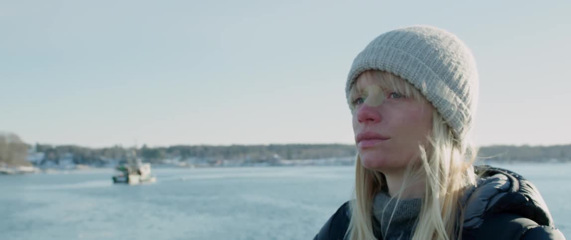 A woman standing on the bow of a ship looks around at the approaching snow covered coast