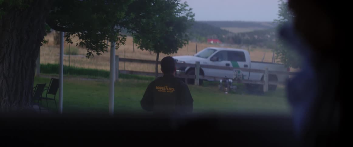 A person pulls the curtains back and looks out the window to see a male walking away towards a truck