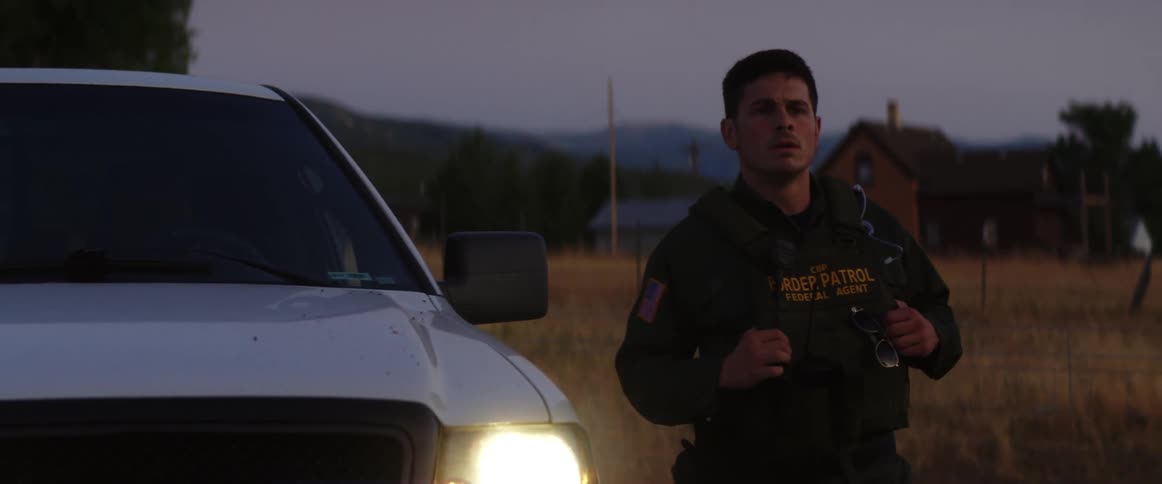 Border patrol agent exits car to stand near vehicle with lit headlights