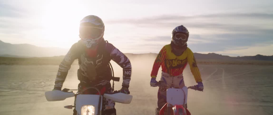 Two people wearing crash helmets drive motorcycles on the desert at sunset