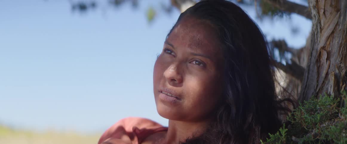 A woman with dirt on her face idles outdoors under the shade of a tree while looking concerned