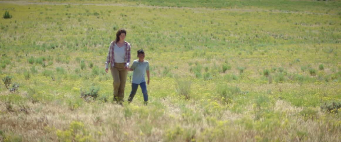A woman and boy walk while holding hands in a field before the boy takes off running