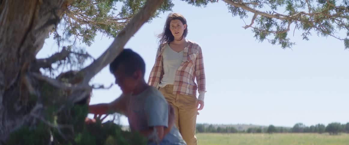 A young boy runs over to hug someone sitting on the ground under a tree with another woman following him
