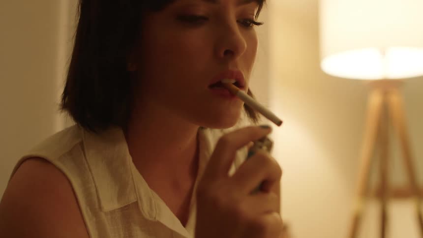Woman with a dark short hair sits on a couch and lights up a cigarette at home