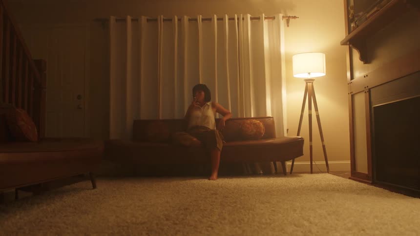 Woman with a dark short hair sitting on a couch by a floor lamp smokes a cigarette in the living room at night