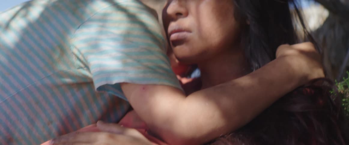 A pregnant woman sits beneath a tree hugging a boy with an anxious expression on her face
