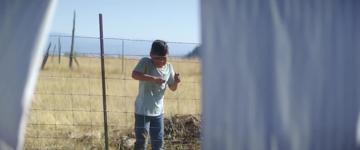 A child shouts and points in an attempt to get the attention of a person hanging laundry on the opposite side of a wire fence before beginning to cry