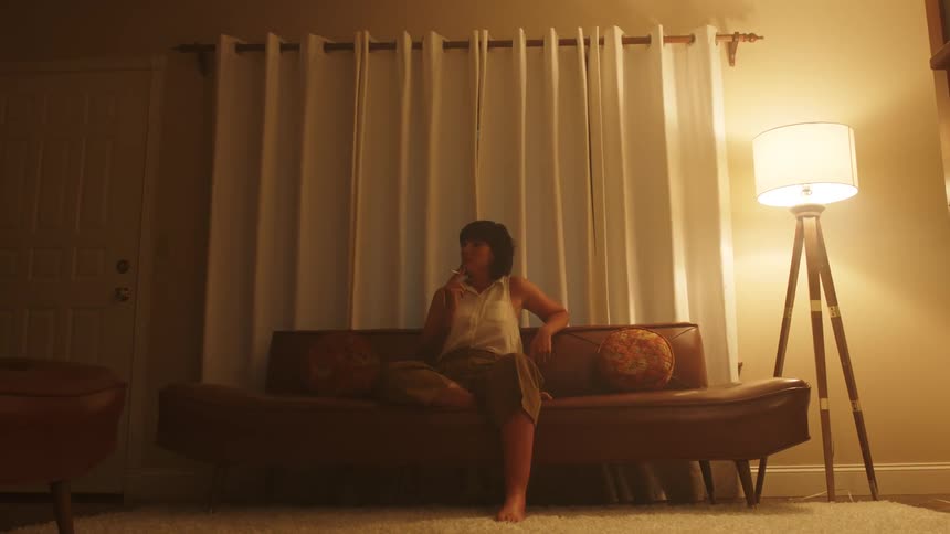 Woman with a dark short hair sits on a couch by a floor lamp smokes a cigarette in a room at night