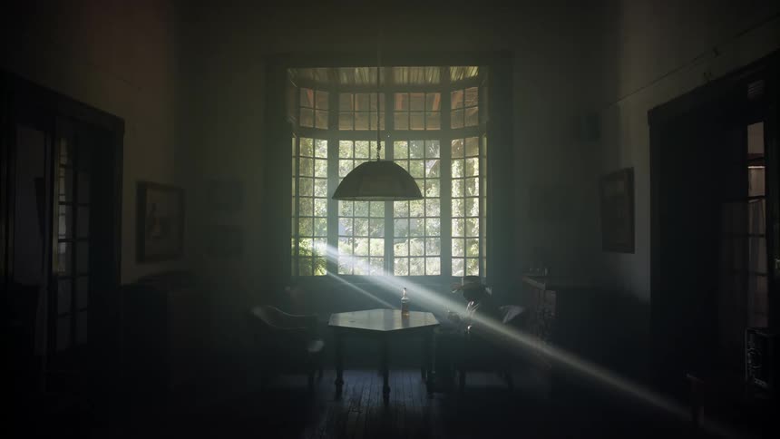 A man sits alone drinking liquor at a table in the dimly lit parlor of a house