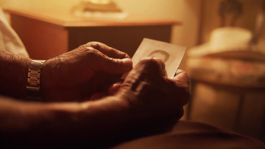 Elderly man sitting on the bed holds and looks at a black and white photograph in a room